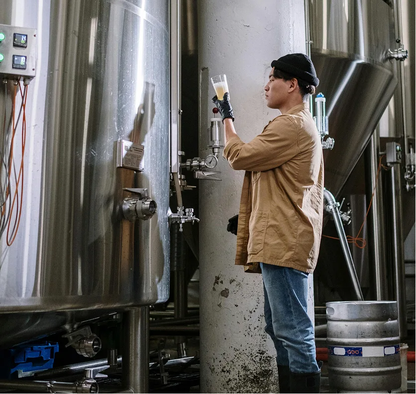 A brewer checking a glass among the fermentation tanks
